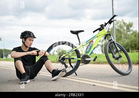 Un giovane e stanco ciclista asiatico seduto per strada con la sua bici a controllare le calorie bruciate e a cavalcare miglia sullo smartwatch. Foto Stock