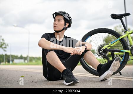 Un giovane e stanco ciclista asiatico in abbigliamento sportivo e un casco da bici siede in strada con la sua bici. sport, esercizio fisico, tempo libero Foto Stock