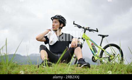 Un giovane asiatico stanco in abbigliamento sportivo riposa sull'erba dopo un lungo giro in bicicletta in montagna. Foto Stock