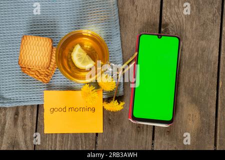 una tazza di tè con i dandeloni al limone e le parole in inglese Good Morning e un telefono con schermo verde Foto Stock