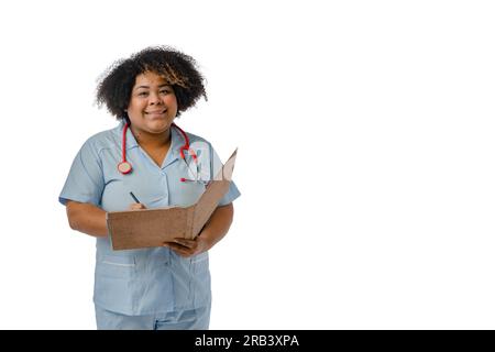 Giovane medico afro-latina che indossa un'uniforme blu e uno stetoscopio rosso sul collo, sorride guardando la macchina fotografica e tenendo un marrone aperto Foto Stock