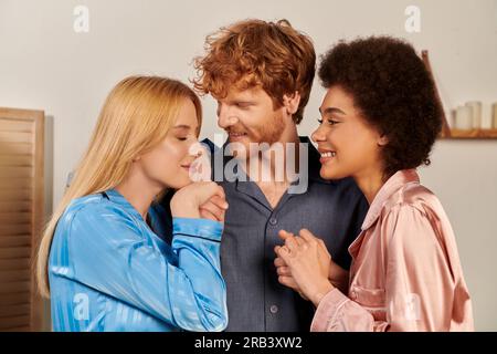 amanti del poliamorio in pigiama, uomo allegro con capelli rossi che abbraccia le donne interrazziali a casa, diversità culturale, partner non tradizionali, libertà in re Foto Stock