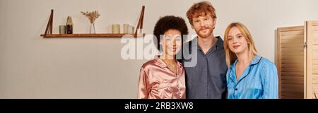 relazioni aperte, uomo felice con i capelli rossi che abbraccia le donne interrazziali a casa, diversità culturale, partner non tradizionali, accettazione e comprensione Foto Stock