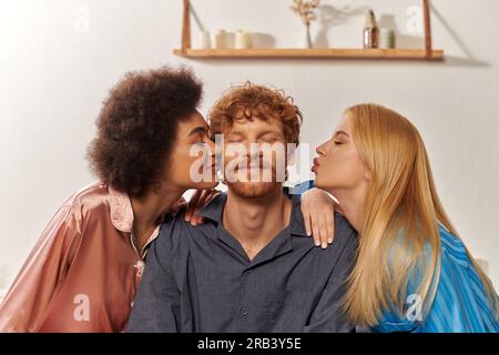 concetto di poligamia, relazione aperta, ritratto di tre adulti, donne multiculturali che baciano l'uomo rosso felice, famiglia poliamorosa in pigiama, culturale Foto Stock