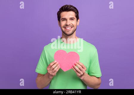 Ritratto di un piacevole ragazzo di buon umore con setole che indossa dita verdi della t-shirt tenendo il cuore di carta isolato su sfondo di colore viola Foto Stock