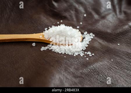 cucchiaino di legno riempito di sale bianco, su pelle marrone, consistenza della pelle, sfondo astratto e delicato Foto Stock