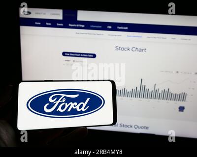 Persona che tiene il cellulare con il logo del produttore automobilistico statunitense Ford Motor Company sullo schermo davanti alla pagina Web aziendale. Concentrarsi sul display del telefono. Foto Stock
