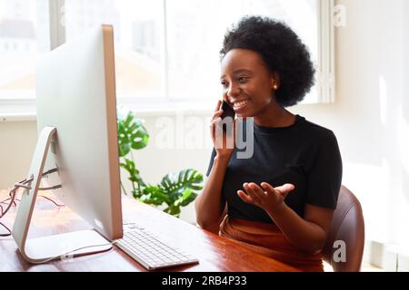 Donna nera sorridente e felice che parla al telefono in ufficio, seduta alla scrivania del PC Foto Stock