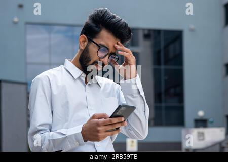 Giovane uomo d'affari ispanico fuori dall'edificio di uffici, uomo in camicia che legge cattive notizie tristi online utilizzando l'app al telefono, uomo depresso deluso infelice dai risultati ottenuti. Foto Stock