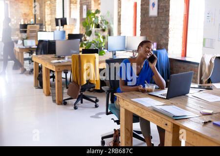Felice donna d'affari afro-americana che parla su smartphone e pranza in ufficio. Affari, cibo, tecnologia e lavoro, inalterati. Foto Stock