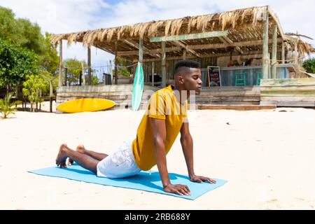 Uomo birazziale concentrato che pratica yoga, si allunga sul tappetino sulla spiaggia soleggiata, inalterato. Posa Cobra, stile di vita sano, benessere, estate e vacanza. Foto Stock