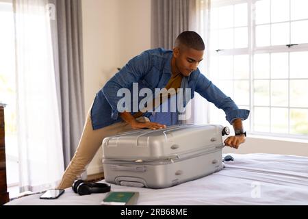 Uomo birazziale che chiude valigia imballata sul letto nella soleggiata camera da letto. Viaggi, vacanze, preparazione, tempo libero e stile di vita inalterati. Foto Stock