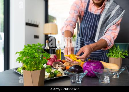 Parte centrale dell'uomo birazziale che indossa il grembiule che prepara il pasto con verdure tritate in cucina. Cibo, cucina, stile di vita sano e vita domestica. Foto Stock