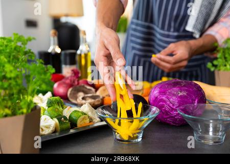 Parte centrale dell'uomo birazziale che indossa il grembiule che prepara il pasto con verdure tritate in cucina. Cibo, cucina, stile di vita sano e vita domestica. Foto Stock