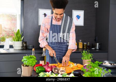 Felice uomo birazziale che indossa il grembiule che prepara il pasto e trita le verdure in una cucina soleggiata. Cibo, cucina, stile di vita sano e vita domestica, inalterati. Foto Stock