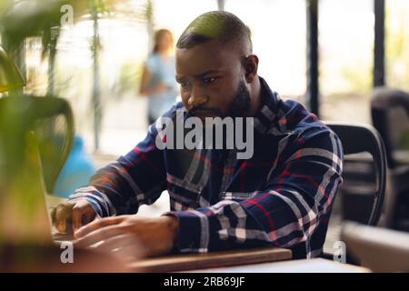 Uomo d'affari afroamericano seduto alla scrivania e che usa il computer portatile in ufficio. Affari occasionali, comunicazione e lavoro, inalterati. Foto Stock