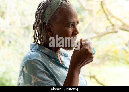 Donna anziana afroamericana premurosa che beve caffè e guarda fuori dalla finestra sul giardino. Stile di vita senior, pensione e vita domestica, unaltere Foto Stock