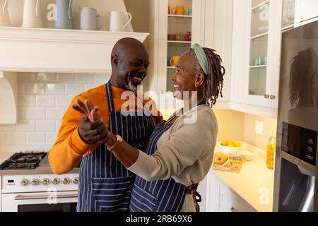 Felice anziano afro-americano che indossa grembiuli e si diverte a ballare insieme in cucina. Stile di vita senior, pensionamento, cucina, insieme e domestico Foto Stock