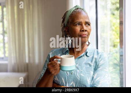 Donna anziana afroamericana premurosa che tiene un caffè e guarda fuori dalla finestra sul giardino. Stile di vita senior, pensione e vita domestica, inalterati Foto Stock