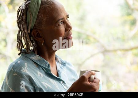 Donna anziana afroamericana felice che tiene un caffè e guarda fuori dalla finestra sul giardino. Stile di vita senior, pensione e vita domestica, inalterati. Foto Stock