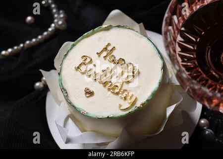 Deliziosa torta decorata con frasi Happy Birthday e bicchiere di vino su sfondo nero Foto Stock