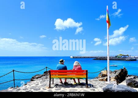 Lanzarote, Isole Canarie, porto di Puerto de Playa Blanca che mostra una coppia anziana seduta al sole con una colomba collettata vicino al molo con moo Foto Stock