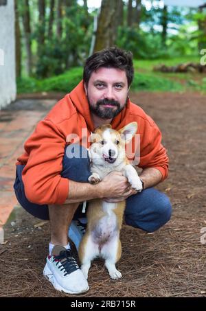 Un giovane uomo barbuto che gioca con il suo cane corgi nella foresta la mattina Foto Stock