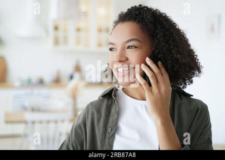 La ragazza sorridente gode di una piacevole telefonata a casa. L'adolescente birazziale riceve buone notizie e parla felicemente con lo smartphone. Foto Stock