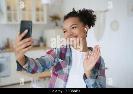 La blogger di razza mista saluta le chiacchiere online via telefono. La signora sorridente in cuffia saluta gli amici, videochiamata a casa. Foto Stock