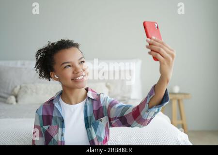 Birazziale teen blogger chatta online tramite videochiamata telefonica, utilizza moderne app a casa. La ragazza sorridente scatta selfie con lo smartphone. Foto Stock