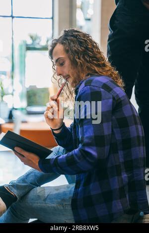 Foto laterale di un giovane uomo dall'aspetto gradevole con lunghi capelli ricci che pensa a una soluzione al suo problema quotidiano al lavoro. Gli ha messo la penna in bocca Foto Stock