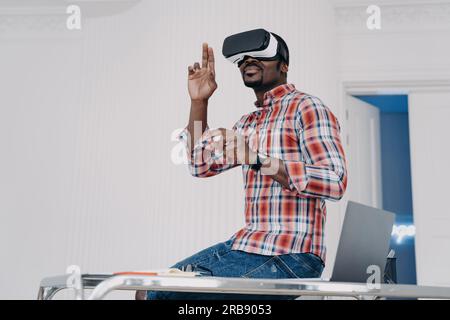 Lo sviluppatore di software afro-americano che indossa occhiali per realtà virtuale, crea app, lavorando nel cyberspazio, seduti sulla scrivania dell'ufficio. Un uomo moderno in realtà virtuale Foto Stock