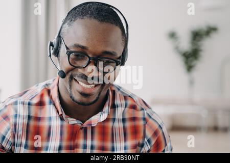 Un uomo afroamericano sorridente, un addetto al servizio di assistenza che indossa cuffie e occhiali, comunica con il cliente. Amichevole operatore di call center nero c Foto Stock
