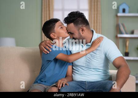 Felice figlio indiano che baciava la fronte del padre mentre sedeva sul divano a casa - concetto di affetto, amore per la famiglia e momento tenero. Foto Stock