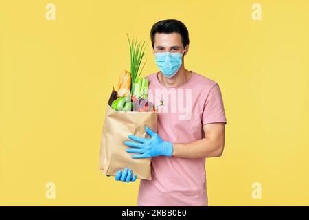 Uomo che tiene in mano una borsa della spesa di carta piena di verdure fresche che indossa maschera e guanti per proteggere le precauzioni contro il coronavirus COVID-19 Foto Stock