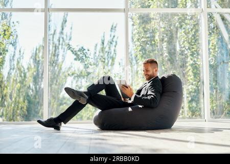 Sorridente uomo d'affari di successo in elegante suite con tablet rilassarsi borsa chair fagiolo con finestre panoramiche di sfondo in moderno creativo ufficio Foto Stock