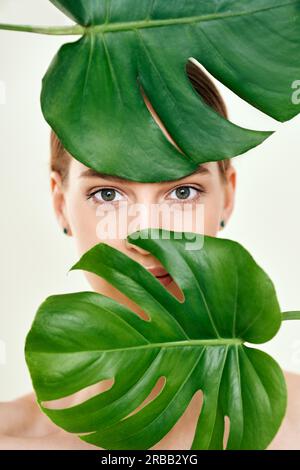 Primo piano ritratto di giovane bella donna con perfetta pelle liscia nascosto in foglie tropicali. Cosmetici naturali, prodotti per la cura della pelle, concetto spa Foto Stock