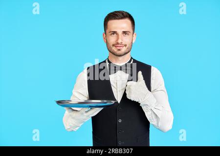 Elegante giovane cameriere che tiene vuoto vassoio d'argento e mostra pollici su segno su sfondo blu. Ristorante, horeca, concetto di successo Foto Stock