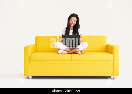 Donna sorridente che usa un computer portatile seduto su un divano giallo su sfondo bianco. Tecnologia, concetto di freelance Foto Stock