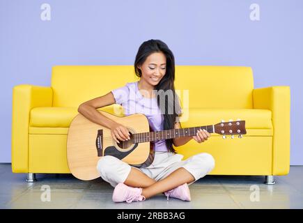 Sorridente donna che suona la chitarra seduta sul divano letto. Concetto creativo, hobby, musica Foto Stock