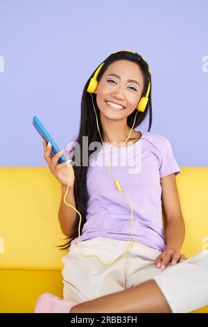 Una ragazza carina che ascolta musica con il cellulare e si rilassa mentre si siede sul divano giallo a casa Foto Stock