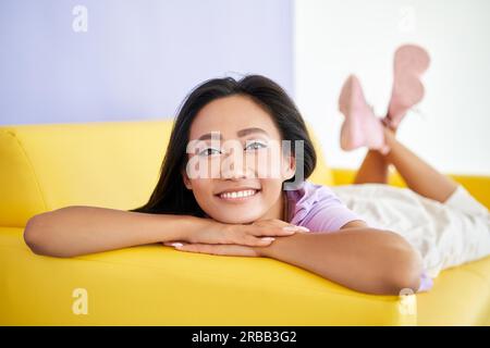 Closeup felice donna asiatica ritratto rilassante a casa sdraiato sul divano giallo. Riposo, bellezza femminile Foto Stock