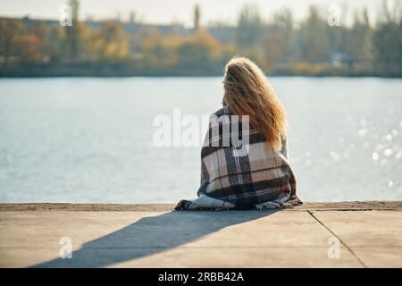 Vista sul retro di una giovane donna che si rilassa da sola lungo un fiume della città avvolta da una coperta di lana e che si gode la natura. Riposo, concetto di viaggio Foto Stock