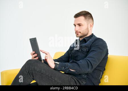 Giovane uomo che guarda il tablet digitale mentre si siede su un divano luminoso. Business, freelance, concetto digitale Foto Stock