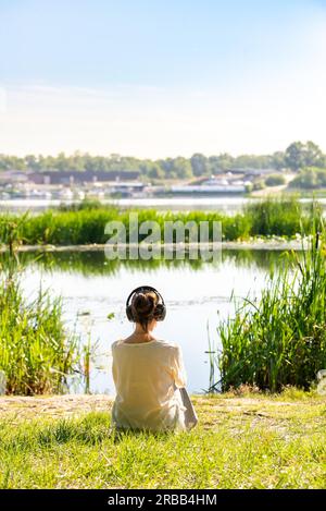 Una donna con le cuffie ascolta musica soffice mentre medita, seduto di fronte al fiume la mattina, subito dopo l'alba Foto Stock