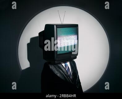 Un uomo con la TV invece della testa. Concetto di zombie mediatico con uomo dipendente dalla televisione in cappotto nero con braccia incrociate in posa sullo studio sotto i riflettori. Foto Stock