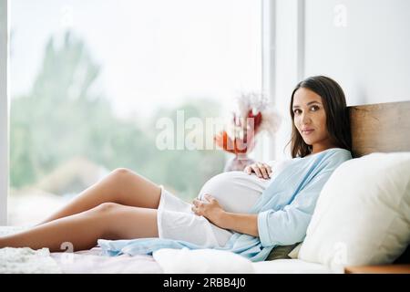 Ritratto di una donna felice incinta sdraiata a letto e toccata la pancia a casa. Maternità, concetto di persone. Tenera foto d'umore della gravidanza Foto Stock