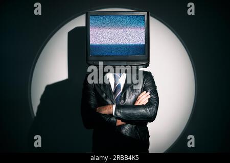 Un uomo con la TV invece della testa. Concetto di zombie mediatico con uomo dipendente dalla televisione in cappotto nero con braccia incrociate in posa sullo studio sotto i riflettori. Foto Stock
