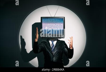 Un uomo con la TV invece della testa. Concetto di zombie mediatico con uomo dipendente dalla televisione in cappotto nero con braccia incrociate in posa sullo studio sotto i riflettori. Foto Stock