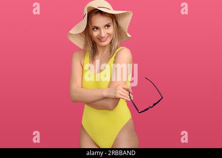 Giovane donna bionda, snella e attraente, indossa un costume da bagno giallo colorato e un cappello da sole in paglia floppy dalle larghe setole che guarda di lato con un felice Foto Stock